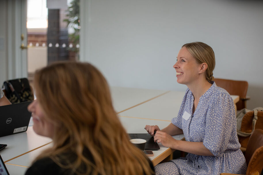 Kaksi ihmistä istuu pöydän ääressä ja katsovat hymyillen eteenpäin. Pöydällä on tietokoneita.