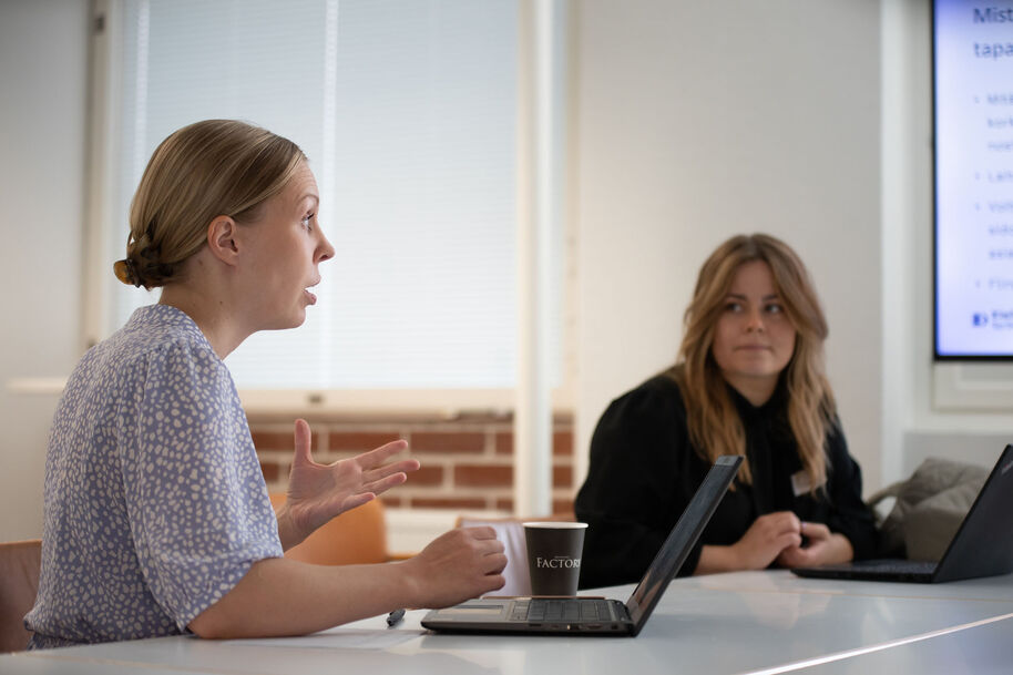 Kaksi naista istuu pöydän ääressä, ja toinen heistä puhuu. Pöydällä on kaksi tietokonetta.