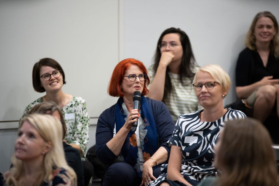 Kaarina Murtola puhumassa mikrofoniin, ympärillä istuu muita kielibuustilaisia.