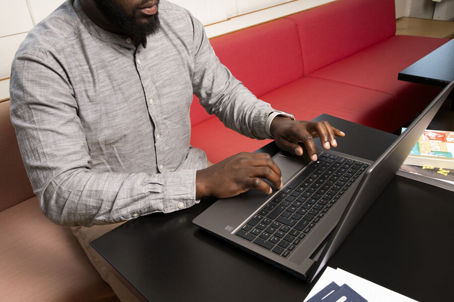 Mies kirjoittaa kannettavalla tietokoneella mustan pöydän ääressä, taustalla punainen sohva.