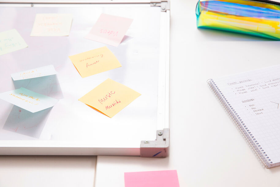 Muistilappuja pöydällä, oikealla käsinkirjoitettu muistikirja ja värikäs penaali.