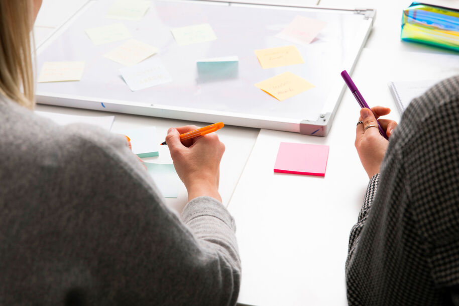 Kaksi ihmistä kirjoittaa muistilappuihin työpöydällä, taustalla valkotaulu ja lisää muistilappuja.