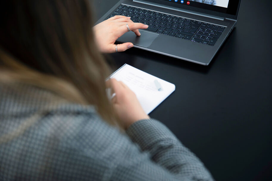 Henkilö kirjoittaa muistikirjaan kynällä ja käyttää kannettavaa tietokonetta mustan pöydän ääressä videopuhelussa.