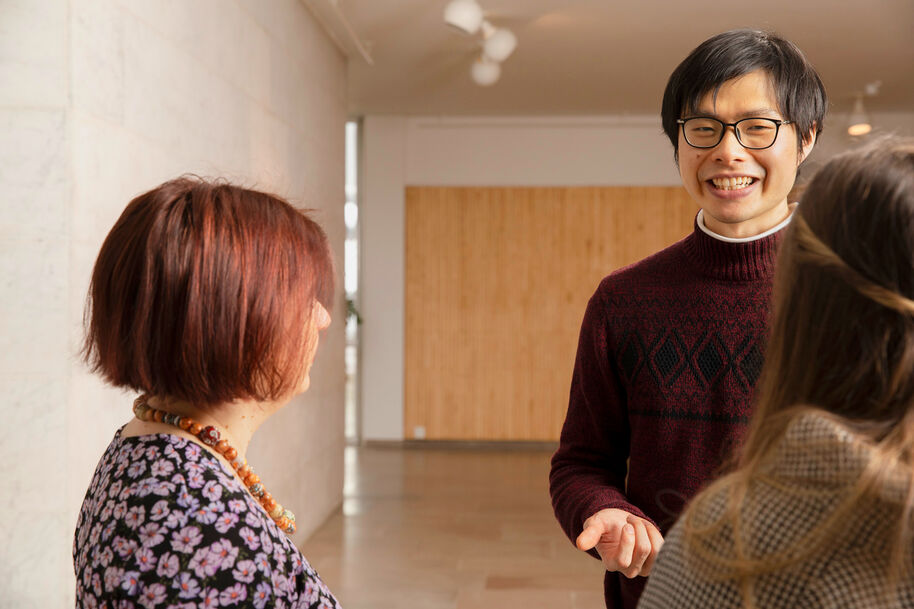 Kolme henkilöä keskustelee käytävällä, jossa on valkoiset ja puupaneelit. Yhdellä on kukkakuvioinen paita ja helmikaulakoru.