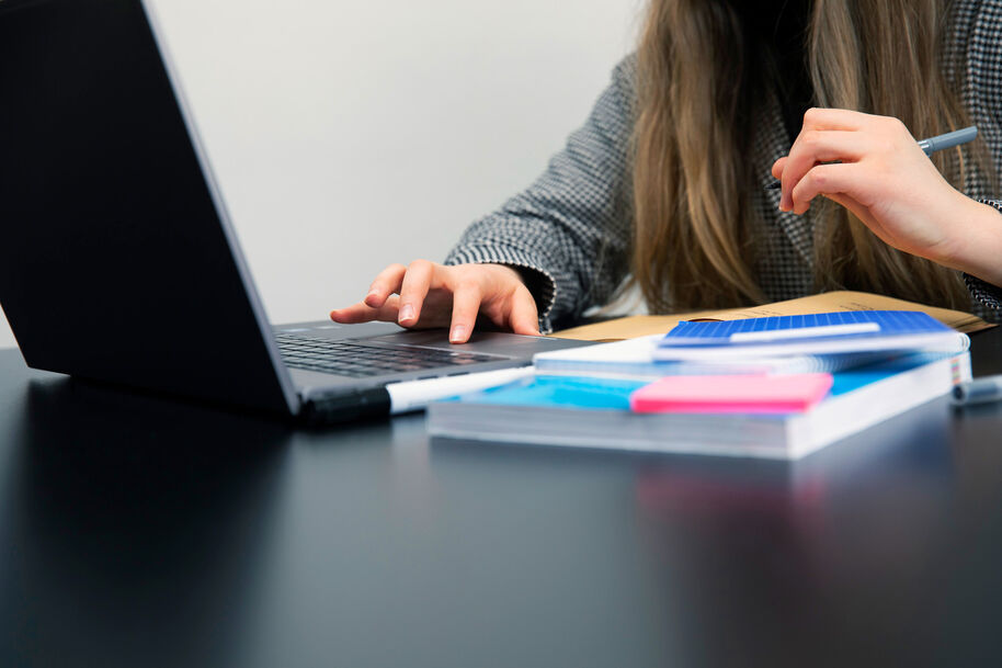 En person arbetar vid en bärbar dator med anteckningsböcker och en penna på bordet.