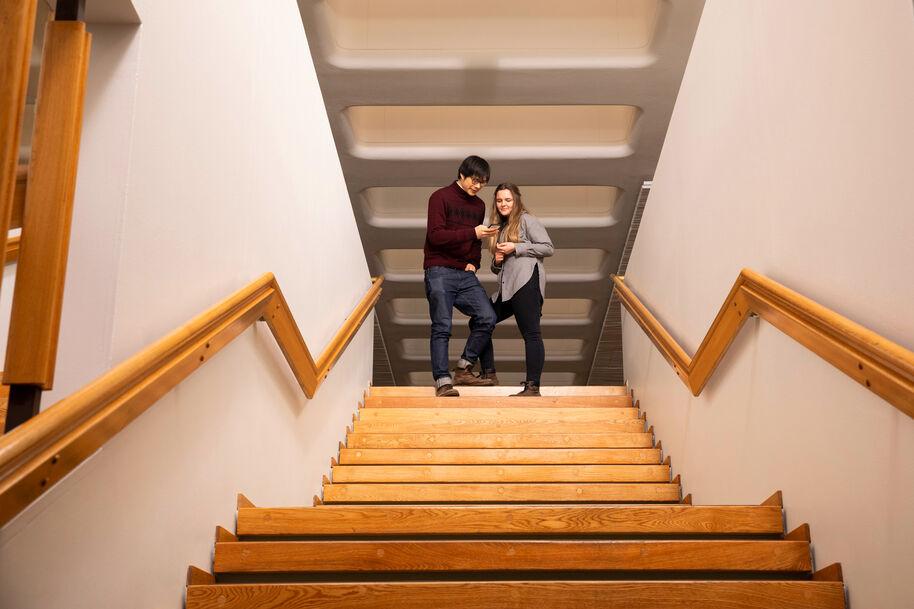 Kaksi henkilöä seisoo puuportaiden yläpäässä ja osoittavat tablettia. Portaissa on puukaiteet.