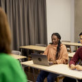 Two students sit in the front row, one looking at the teacher and the other at a computer.