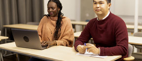 Opiskelija katsoo hymyillen eteenpäin, ja hänen vieressä istuva opiskelija katsoo tietokonetta.