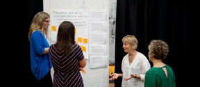 Four persons are talking next to a wall. There is a large sheet of paper with notes and notepads on the wall.