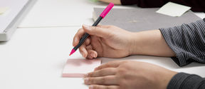 Kädet, jotka kirjoittamassa kynällä muistilapulle. 