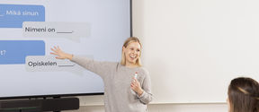 Opettaja seisoo luokan edessä ja osoittaa taululle, jossa on suomen kielen keskusteluharjoitus. 