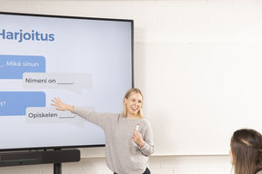 Opettaja seisoo luokan edessä ja osoittaa taululle, jossa on suomen kielen keskusteluharjoitus. 
