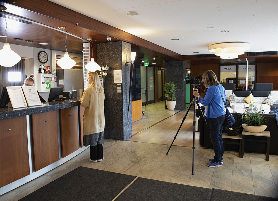Kuva on hotellin aulasta. Asiakas seisoo vastaanottotiskin edessä ja kuvaaja seisoo kameran takana aulassa ja kuvaa tilannetta. 