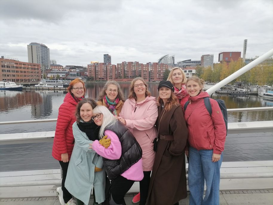 8 hymyilevää henkilöä seisomassa Tampereen Laukonsillalla, taustalla kaupunkimaisema